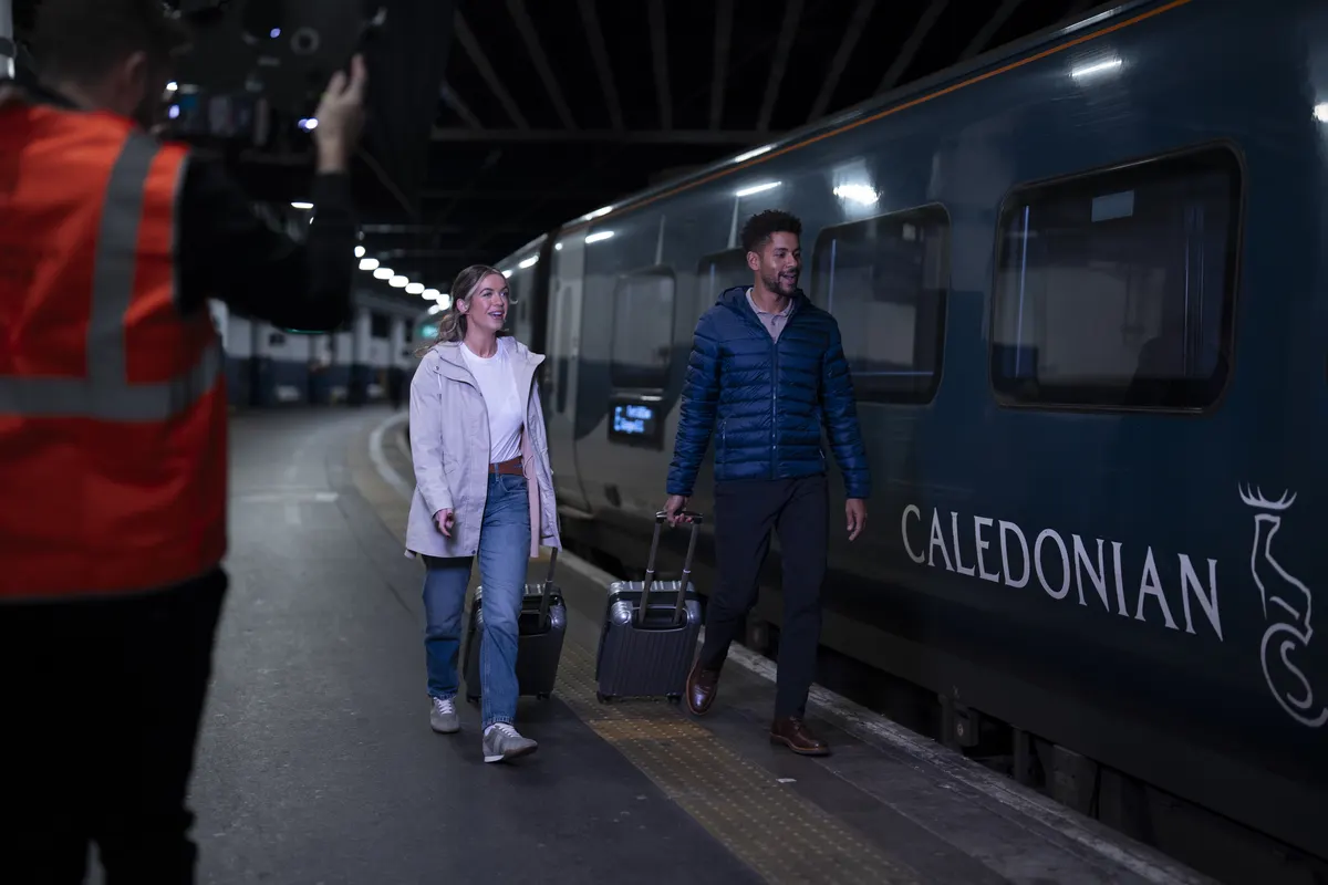 Camera operator filming two travellers beside a Caledonian Sleeper train on a platform