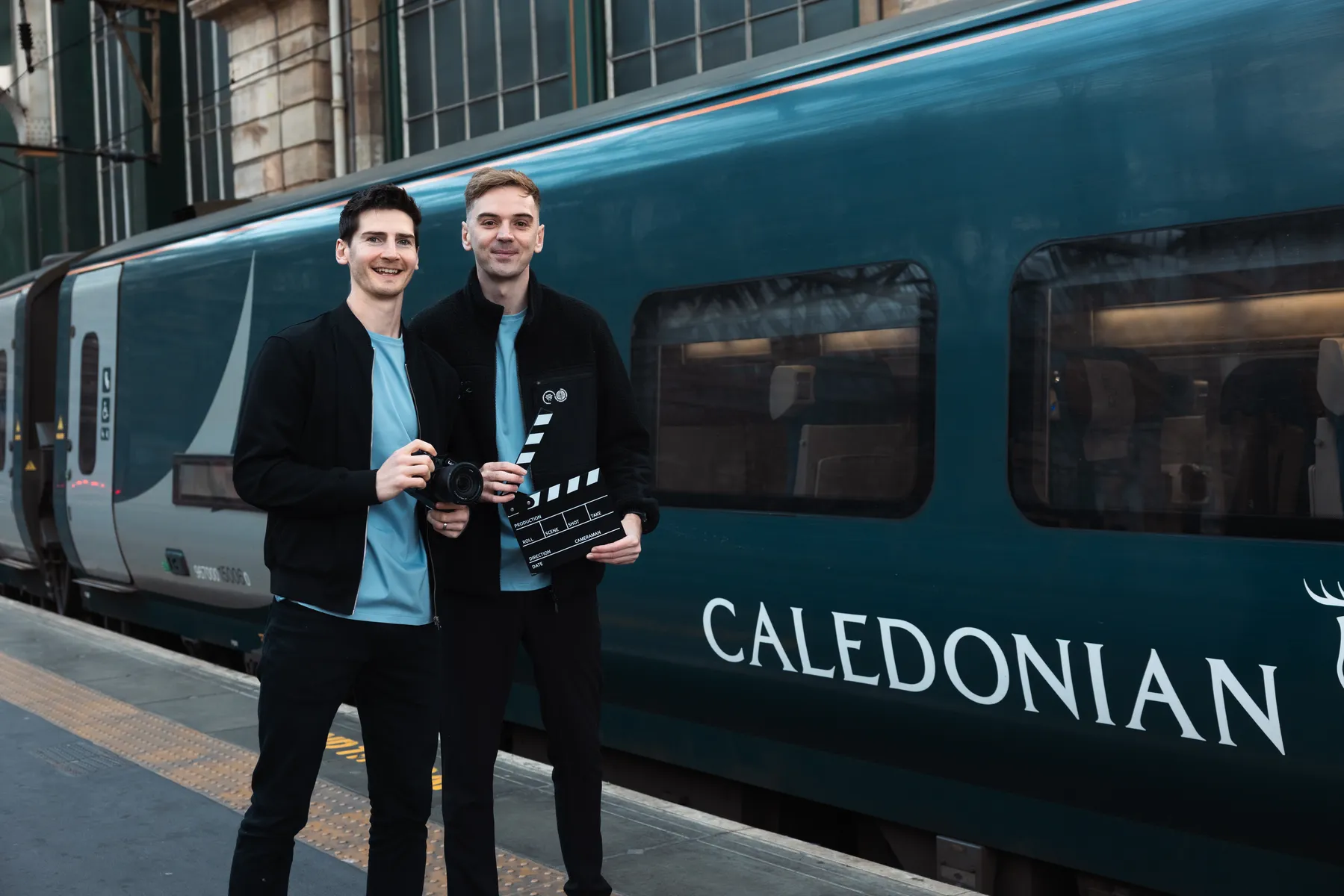 Two film crew members with a camera and clapperboard beside the Caledonian Sleeper train
